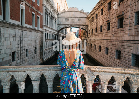 Vista posteriore della donna che indossa hat permanente, mentre dalla ringhiera sul ponte in mezzo a edifici in città Foto Stock