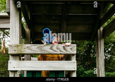 Ragazza guardando attraverso il telescopio mentre si sta in piedi sul legno gioco all'aperto Attrezzature al parco giochi Foto Stock