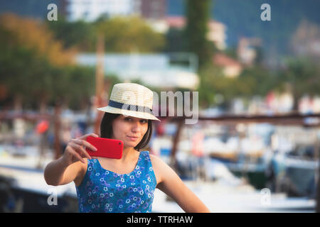 Donna sorridente indossando hat tenendo selfie mentre permanente al porto in città Foto Stock