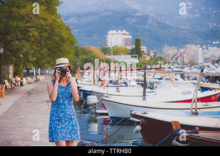 Donna che indossa hat fotografare con la fotocamera mentre si sta in piedi in porto in città Foto Stock