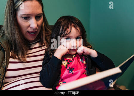 Donna incinta con figlia libro lettura stando seduti a casa Foto Stock