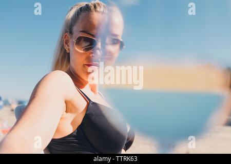 Ritratto di donna che indossa gli occhiali da sole e bikini in spiaggia con bicchiere in primo piano Foto Stock