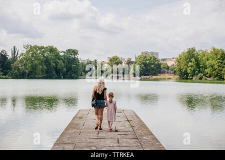 Vista posteriore della madre con la figlia camminando sul molo sul lago contro il cielo nuvoloso in città Foto Stock