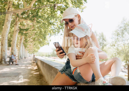 Madre utilizzando smart phone mentre è seduto con la figlia sulla parete di ritegno in posizione di parcheggio Foto Stock