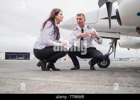 Ingegnere maschio spiegando a femmina partecipante mentre accovacciato in aereo sulla pista di aeroporto contro il cielo nuvoloso Foto Stock