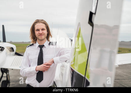 Ritratto di fiducioso pilota maschio in piedi in aereo contro il cielo nuvoloso sulla pista di aeroporto Foto Stock