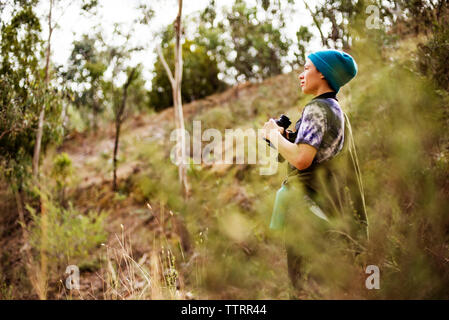 Vista laterale della donna che guarda lontano mentre si tiene il binocolo in foresta Foto Stock