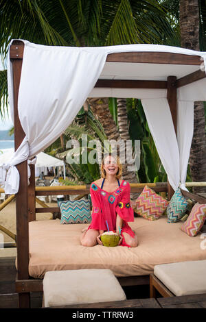 Ritratto di donna felice con noce di cocco in ginocchio sul letto a baldacchino a beach Foto Stock