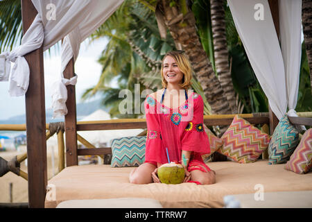 Donna felice con noce di cocco in ginocchio sul letto a baldacchino a beach Foto Stock