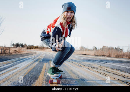 Ritratto di donna giocoso gesti segno di pace mentre lo skateboard su strada durante il periodo invernale Foto Stock