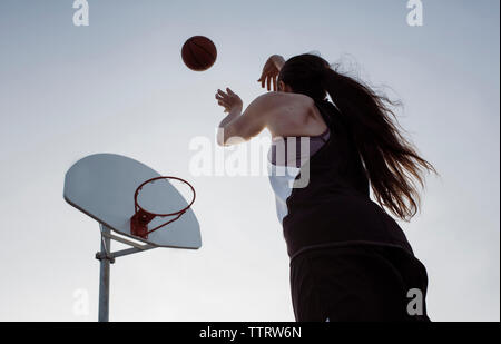 Basso angolo di visione della donna giocare a basket mentre in piedi contro il cielo chiaro durante il tramonto Foto Stock