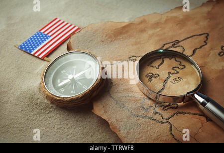 Mappa vecchia con la bussola, bandiera americana e alla lente di ingrandimento sulla sabbia. Felice il Columbus day concept Foto Stock