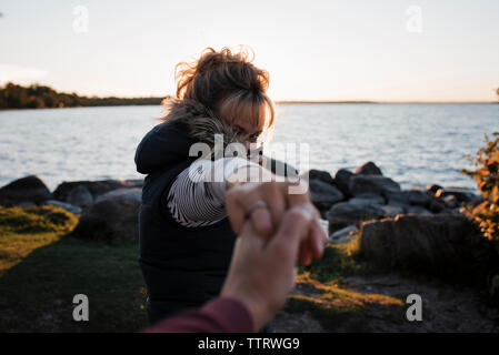 Immagine ritagliata del marito moglie tenendo la mano in piedi sul lungolago contro il cielo chiaro durante il tramonto Foto Stock