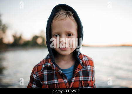 Ritratto di carino ragazzo in maglia con cappuccio in piedi dal lago contro il cielo chiaro durante il tramonto Foto Stock