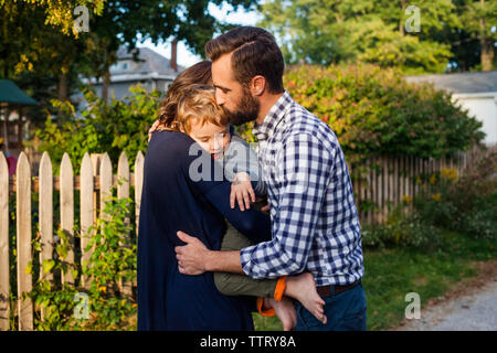 Vista laterale dei genitori abbracciando e baciando figlio mentre in piedi contro piante in cantiere Foto Stock