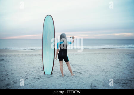 Vista posteriore del surfista femmina fotografando attraverso smart phone mentre in piedi da tavola da surf su Delray Beach Foto Stock