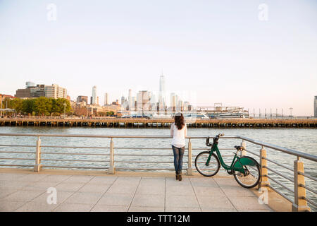 Vista posteriore della donna con la bicicletta alla ricerca in vista della città mentre si sta in piedi sul punto di osservazione Foto Stock