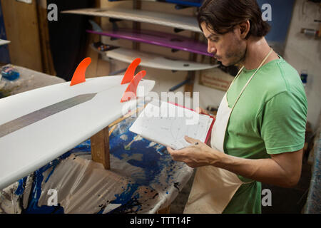 Lavoratore di sesso maschile guardando le tavole da surf schemi nel libro mentre in piedi in officina Foto Stock