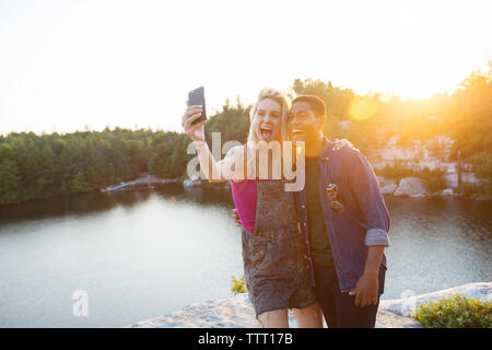 Allegro amici prendendo selfie attraverso la telecamera dal lago durante il tramonto Foto Stock