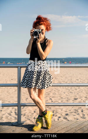 Giovane donna fotografare attraverso la telecamera mentre in piedi dalla ringhiera a beach Foto Stock