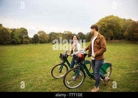 Coppia felice di parlare mentre si cammina con le biciclette sul campo erboso Foto Stock