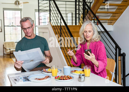 Uomo che legge il giornale mentre donna utilizzando smart phone al tavolo da pranzo Foto Stock