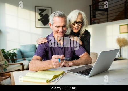 Donna sorridente guardando il computer portatile mentre uomo con carta di credito in salotto Foto Stock