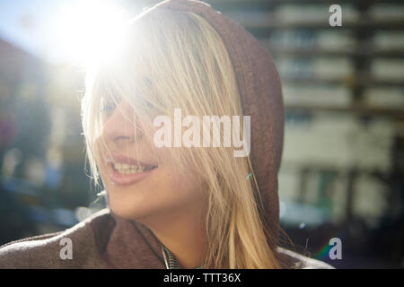 Close-up di felice donna con capelli biondi indossa maglietta con cappuccio mentre in piedi in città durante la giornata di sole Foto Stock