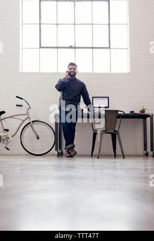 Imprenditore sorridente parlando al telefono mentre è appoggiata dalla tavola contro la finestra Foto Stock