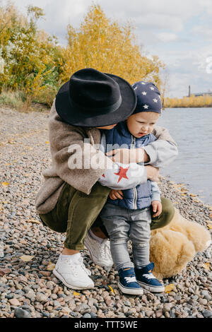 Madre abbracciando carino figlio mentre accovacciato sui ciottoli sul lungolago contro sky nel parco durante l'autunno Foto Stock