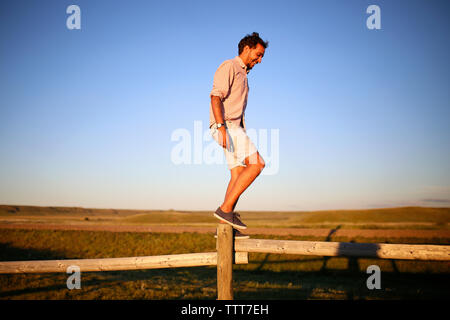 Lunghezza completa di uomo felice bilanciamento sulla recinzione di legno contro il cielo blu e chiaro durante il tramonto Foto Stock