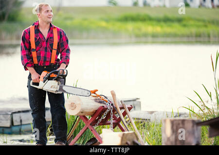 Uomo sorridente con chainsaw registro di taglio a riverbank Foto Stock