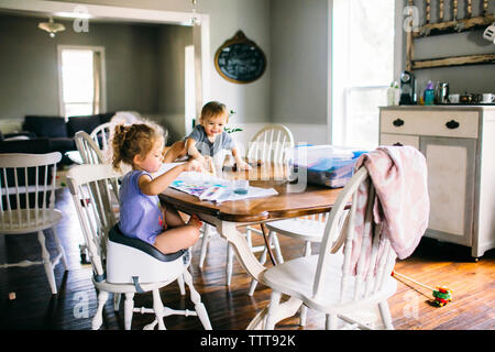 Suor pittura mentre il fratello seduto sul tavolo da pranzo a casa Foto Stock