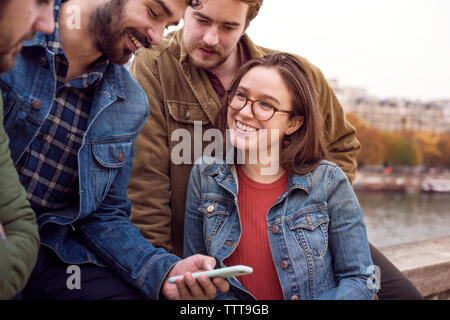 Felice l'uomo utilizzando smart phone con gli amici mentre è seduto sulla parete di ritegno Foto Stock