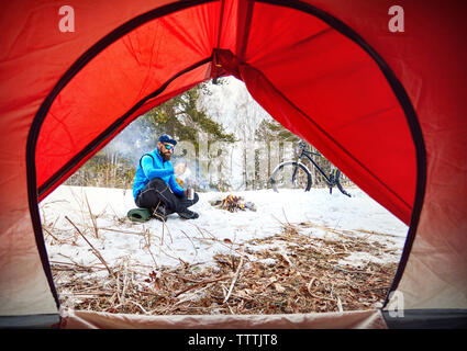 Turismo maschio il caffè seduti al campeggio nel bosco visto attraverso la tenda Foto Stock