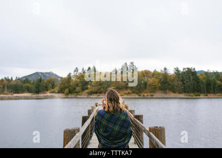 Vista posteriore della donna avvolta in una coperta a piedi sul molo sul lago contro il cielo chiaro Foto Stock