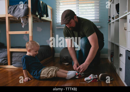 Padre mettendo le scarpe sul figlio mentre inginocchiato a casa Foto Stock