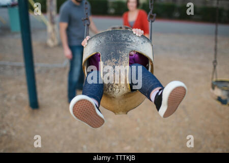 Figlia swinging presso il parco giochi con i genitori in piedi in background Foto Stock