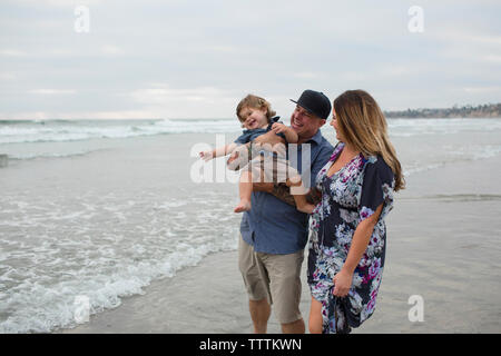 Felice madre guardando il figlio essendo portati dal padre sulla riva contro sky Foto Stock