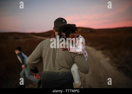 Vista posteriore del padre che porta la figlia mentre si cammina sulla strada sterrata durante il tramonto Foto Stock