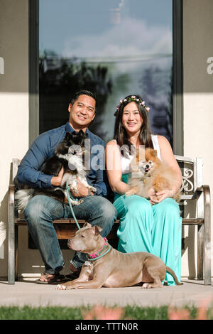 Ritratto di fiducioso sorridente giovane con cani seduti sulla panchina contro house Foto Stock