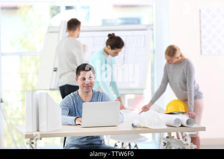 Giovane maschio ingegnere di progetto di pianificazione su computer e altri ingegneri con tavola da disegno in office Foto Stock