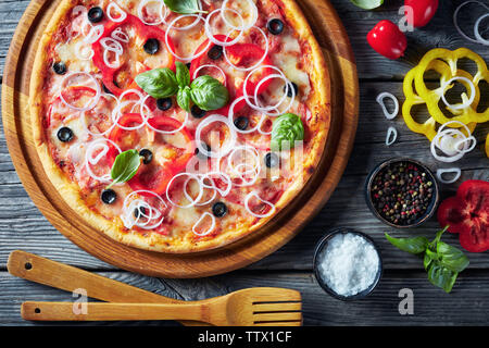Pizza con pomodoro, formaggio, cipolla, prosciutto, olive e peperone anelli su di un tagliere di legno su un livello di grigio tavolo rustico con ingredienti, vista da ab Foto Stock