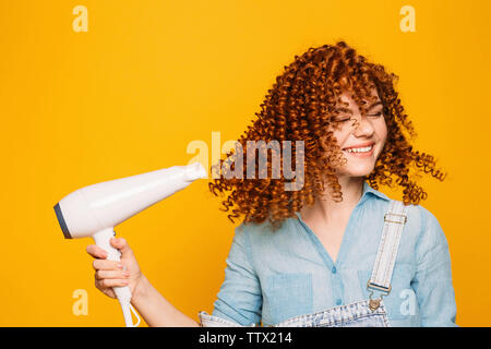 Ricci i capelli rossi donna utilizzando asciugacapelli su sfondo giallo. Fare ricci perfetti Foto Stock