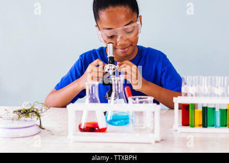 Gioiosa nero bella carino ragazza intelligente in occhiali protettivi seduta nella scuola laboratorio chimico e cercando di microscopio Foto Stock