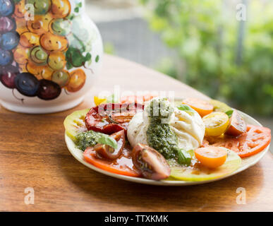 Fresh multi colorata insalata di pomodoro con mozzarella di bufala e il verde del basilico Pesto, su un tavolo esterno con piante in background Foto Stock