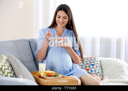 Giovane donna incinta con il latte e la colazione sul divano Foto Stock