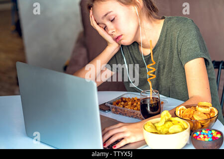 Ragazza lavora a un computer e si mangia il fast food. Cibo malsano: chips, cracker, candy, waffle, cola. Il cibo spazzatura, concetto. Foto Stock