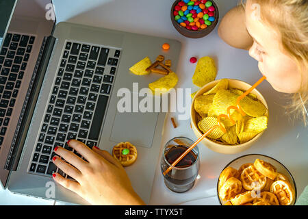 Ragazza lavora a un computer e si mangia il fast food. Cibo malsano: chips, cracker, candy, waffle, cola. Il cibo spazzatura, concetto. Foto Stock