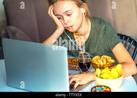 Ragazza lavora a un computer e si mangia il fast food. Cibo malsano: chips, cracker, candy, waffle, cola. Il cibo spazzatura, concetto. Foto Stock